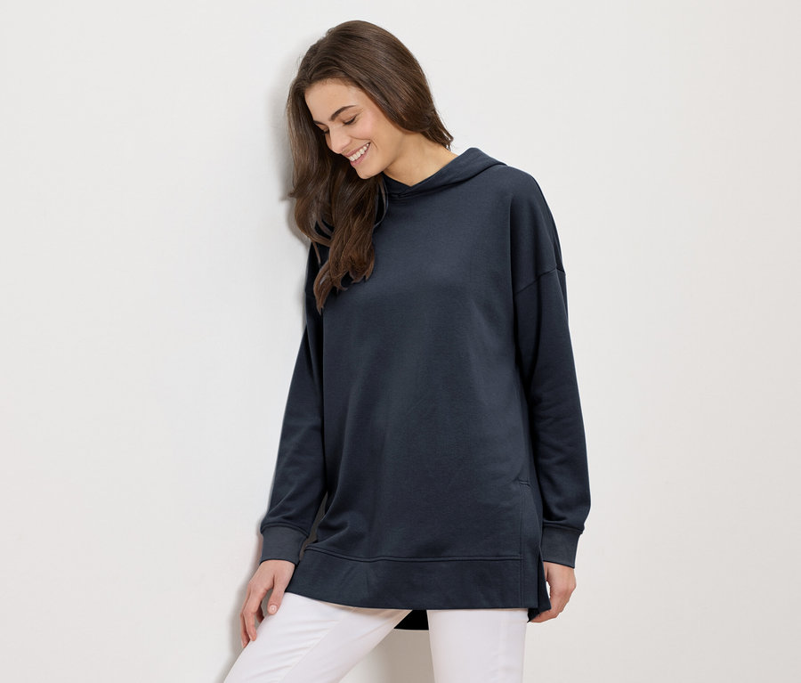 Une femme aux longs cheveux bruns portant un long sweat à capuche bleu marine et un pantalon blanc sourit et s'appuie contre un mur blanc.