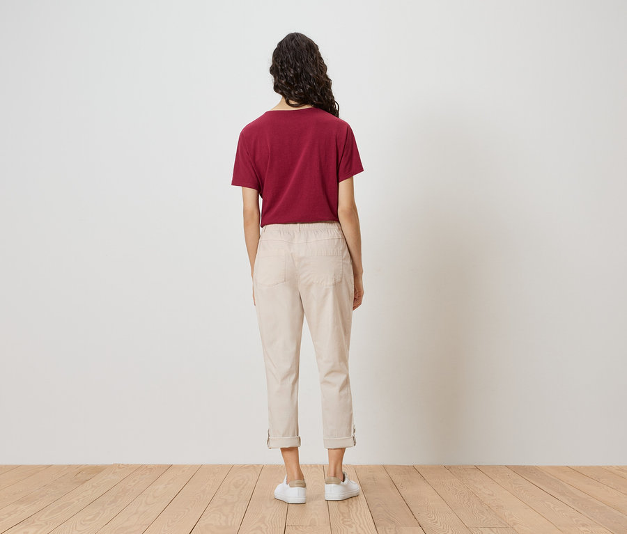 Une femme, vue de dos, porte un t-shirt bordeaux, un pantalon beige et des chaussures blanches.