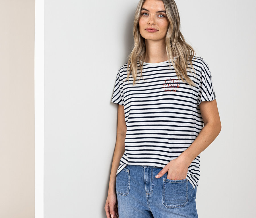 Une femme blonde portant un t-shirt Street One bleu marine et blanc et un jean bleu est appuyée contre un mur.