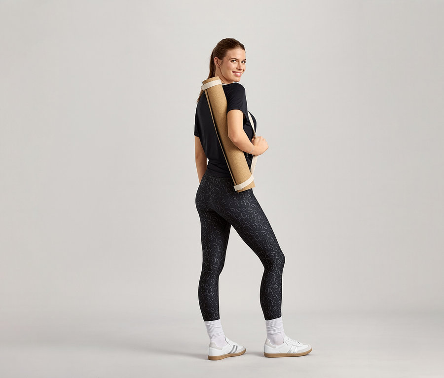 Femme souriante avec un tapis de yoga sur l'épaule, portant un t-shirt de sport noir.