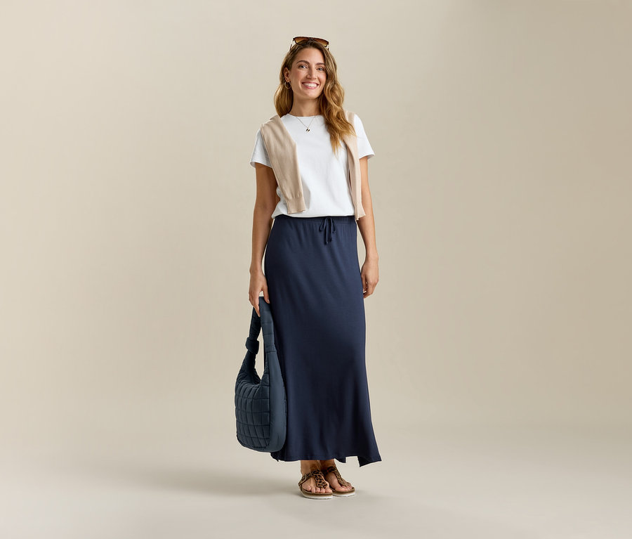 Femme souriante portant un t-shirt blanc et une jupe longue en jersey, tenant un sac bleu.