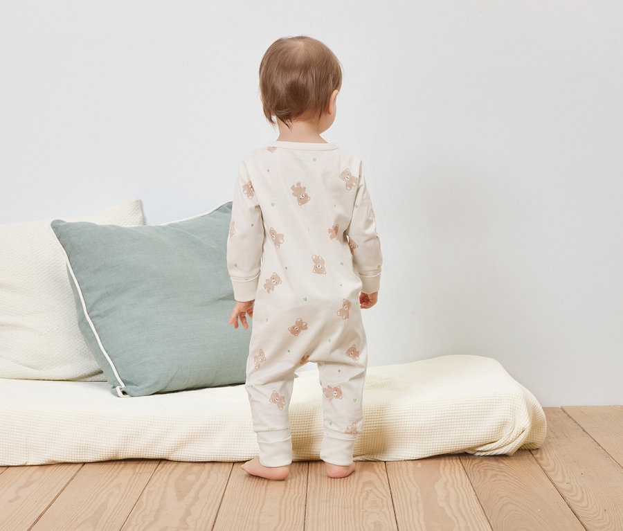 Un enfant se tient dos à nous sur un plancher en bois, vêtu d'une combinaison beige à motifs d'ours.