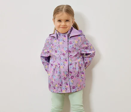 Une jeune fille blonde porte un imperméable violet à motifs floraux et un pantalon vert clair devant un mur blanc.