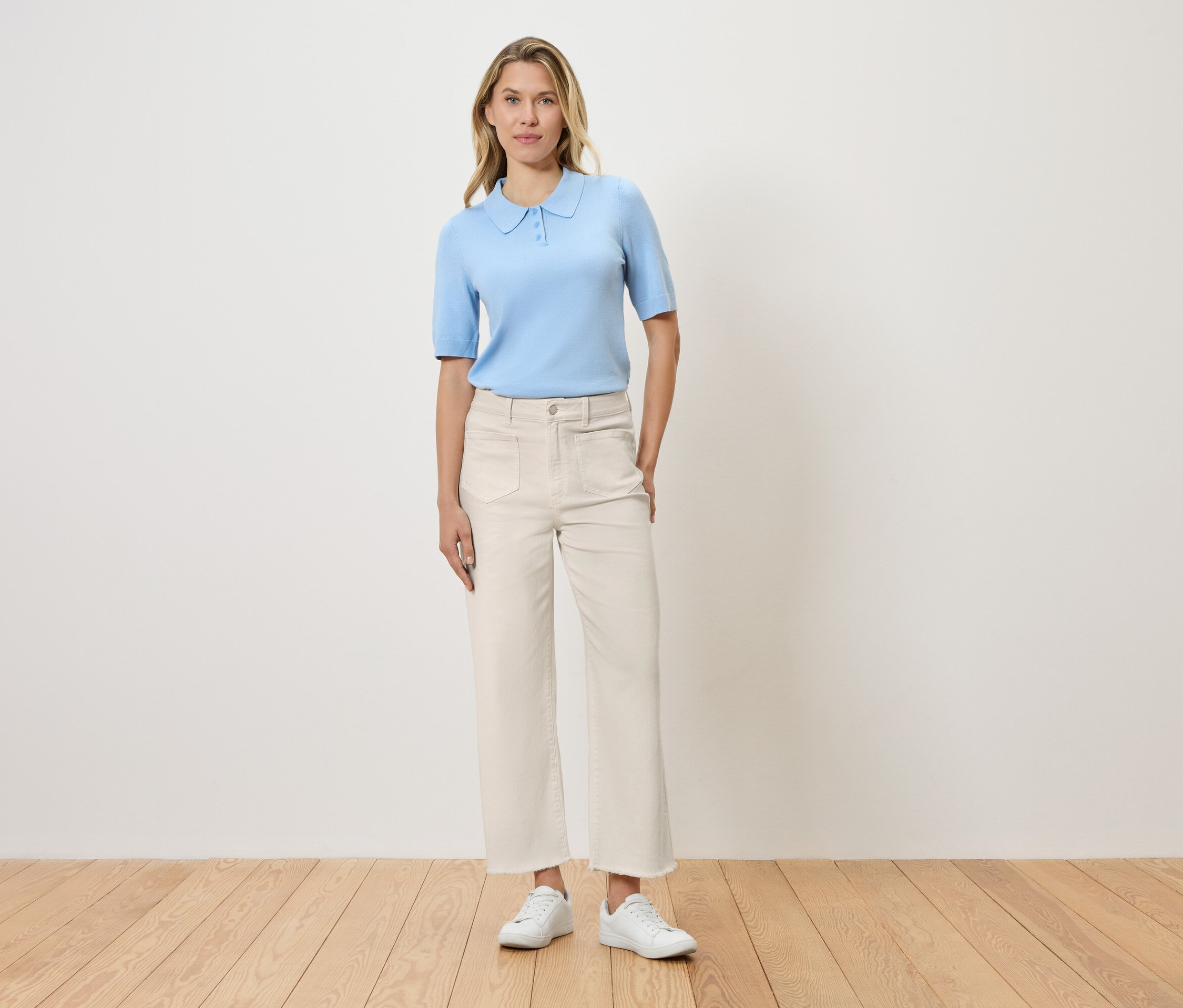 Une femme debout devant un mur, portant un polo bleu clair, un pantalon blanc et des chaussures blanches.