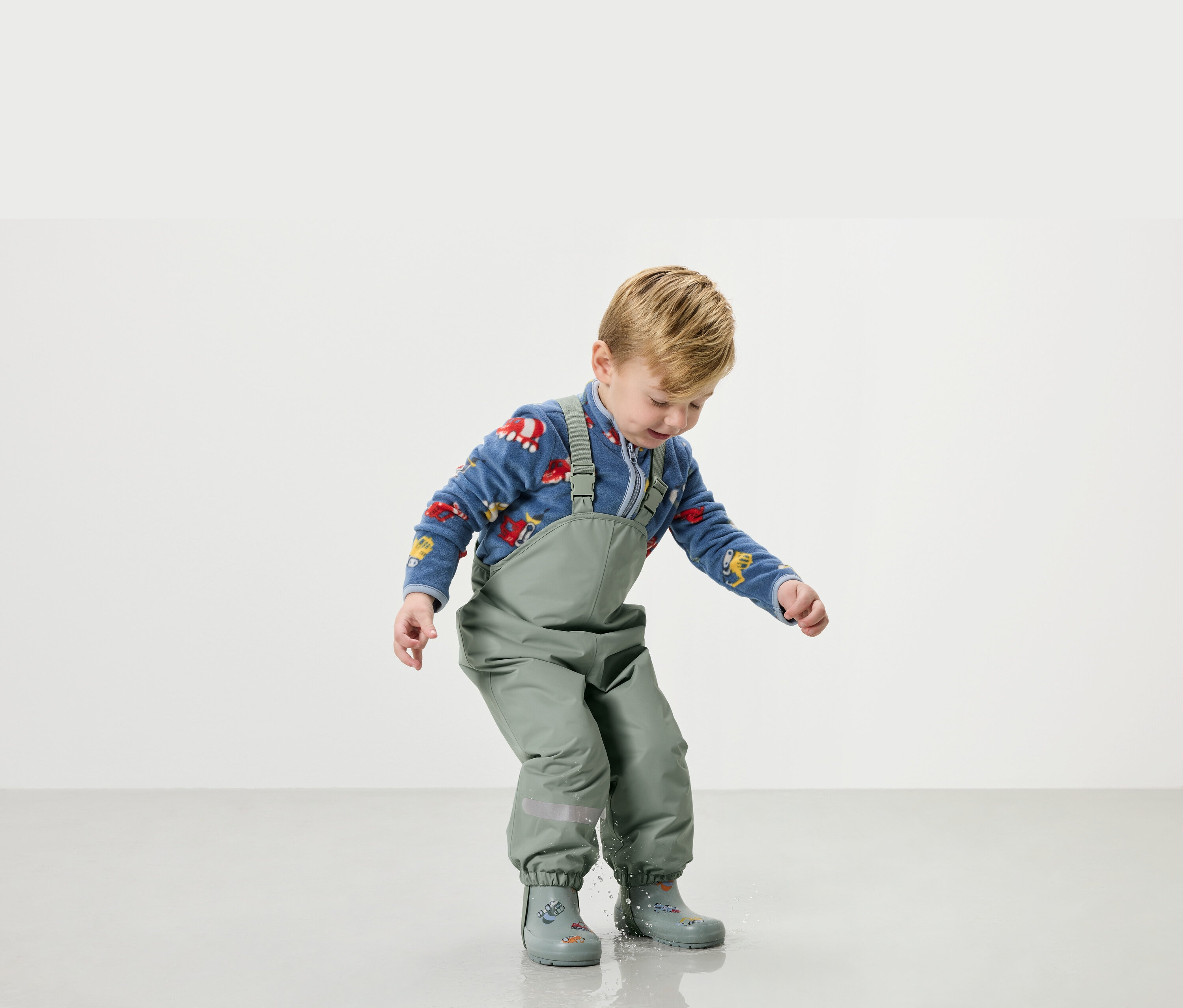 Un jeune garçon joue dans l'eau avec un habit de pluie vert et des bottes en caoutchouc.