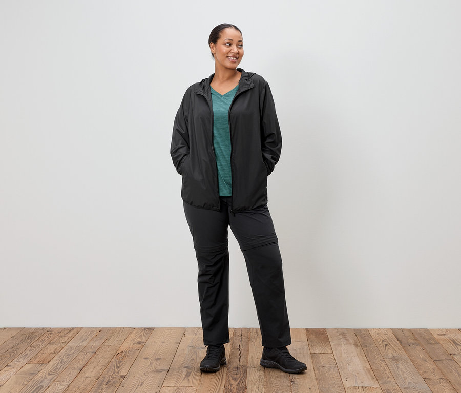 Une femme se tient debout, les mains dans les poches, portant un imperméable noir, un haut vert et un pantalon noir, devant un mur blanc.