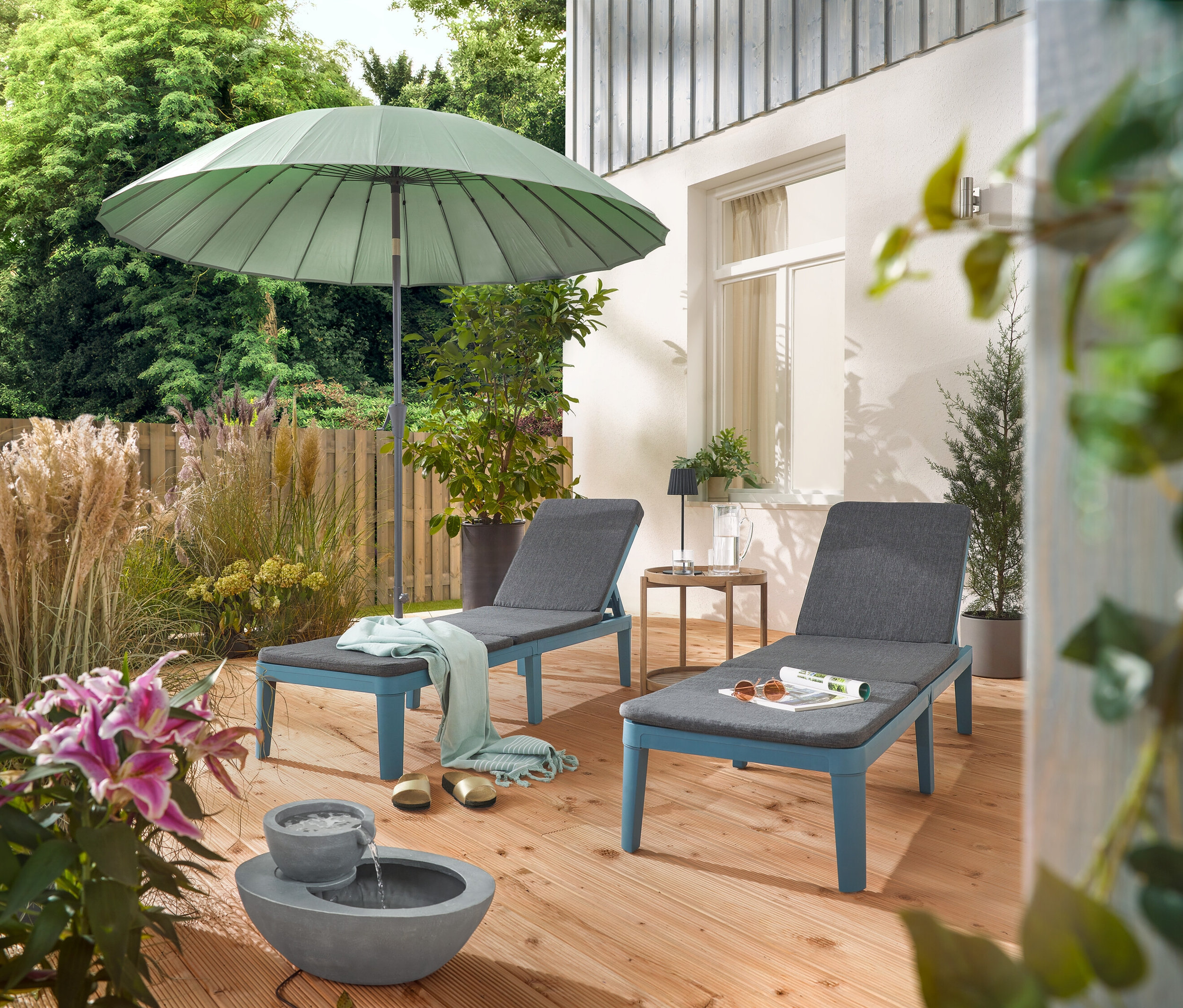 Sur une terrasse, deux chaises longues sont placées sous un parasol Leco Shanghai gris clair et à côté d'une fontaine de jardin solaire.