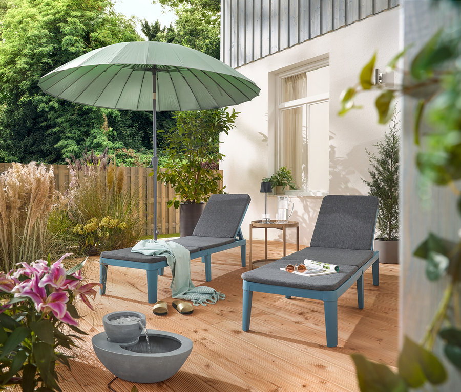 Sur une terrasse, deux chaises longues sont placées sous un parasol Leco Shanghai gris clair et à côté d'une fontaine de jardin solaire.