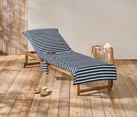 Une chaise longue avec une housse à rayures bleues et blanches se trouve sur un plancher en bois, à côté d'un sac et des sandales.