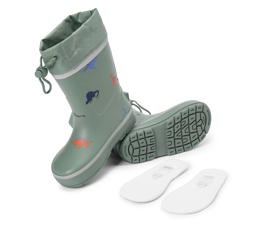 Une paire de bottes en caoutchouc vertes à motif de dinosaures et deux semelles intérieures blanches sont posées sur un fond blanc.