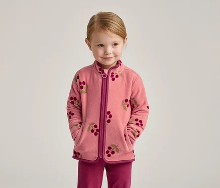 Une fillette blonde porte une veste en polaire rose à motif cerises et un pantalon rose.