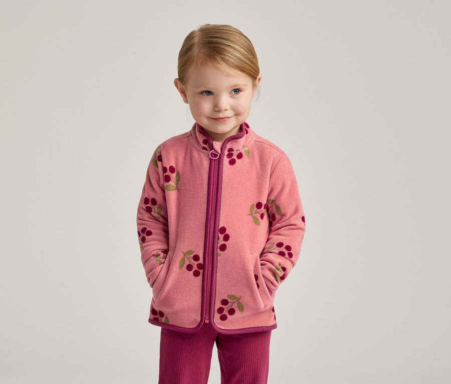 Une fillette blonde porte une veste en polaire rose à motif cerises et un pantalon rose.