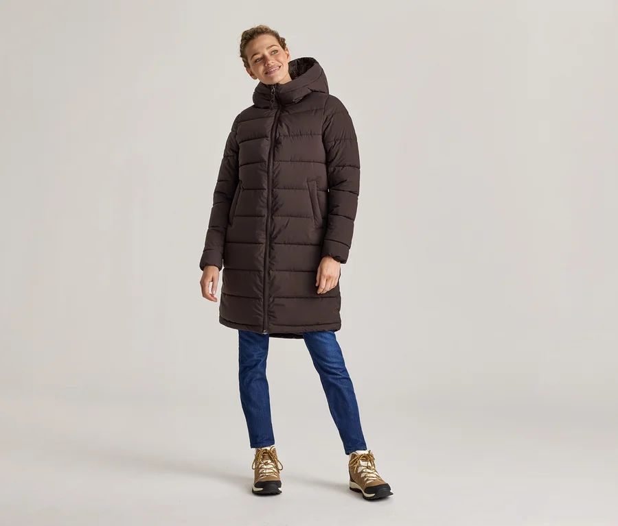 Une femme en manteau bouffant long marron, jean et chaussures de randonnée se tient devant un fond clair.