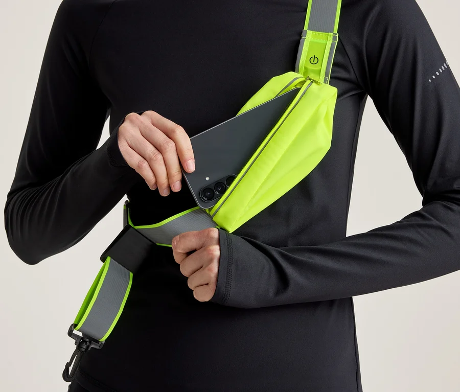 Personne en haut à manches longues noir insérant un smartphone noir dans un sac banane jaune fluo avec un détail réfléchissant.