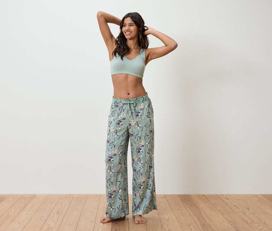 Femme portant un soutien-gorge de sport vert clair et un pantalon de pyjama à fleurs, debout devant un mur.