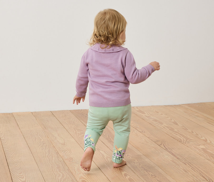 Un jeune enfant aux cheveux blonds se tient pieds nus sur un plancher en bois, portant un pull violet et un pantalon vert clair avec des broderies florales.