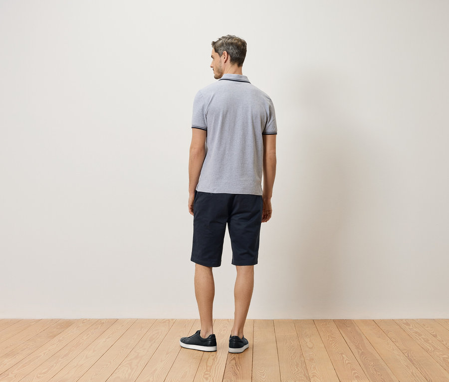Homme de dos, portant un polo gris, un short bleu et des chaussures noires.