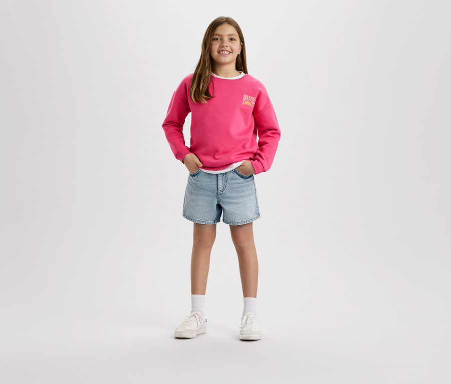 Une fille portant un pull rose et un short en jean se tient devant un fond blanc.