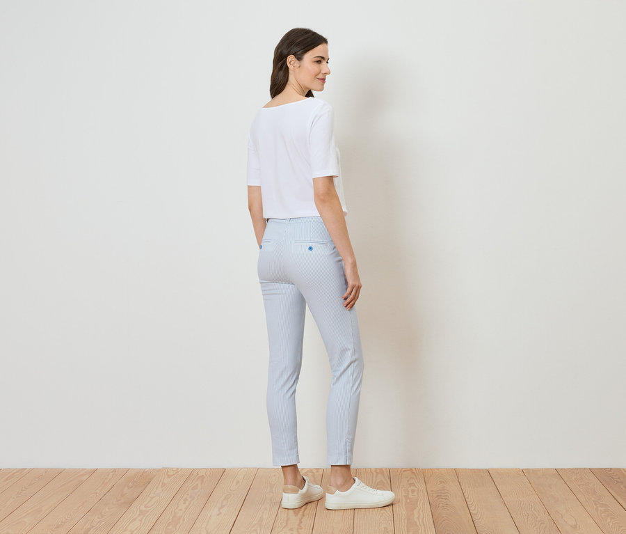 Une femme se tient dans une pièce lumineuse, portant un t-shirt blanc, un pantalon rayé bleu clair et des chaussures blanches.