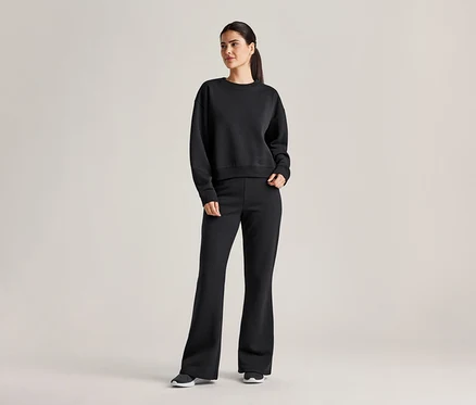 Une femme portant une queue de cheval porte un sweat-shirt noir et un pantalon évasé avec des baskets sur un fond gris clair.