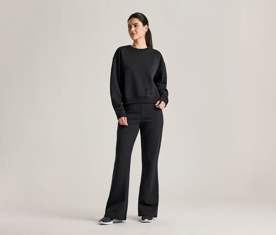 Une femme portant une queue de cheval porte un sweat-shirt noir et un pantalon évasé avec des baskets sur un fond gris clair.