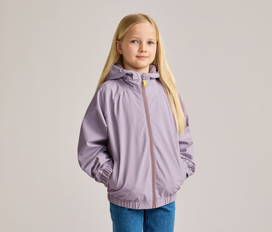 Une fille blonde, en jean et veste à capuche violette, les mains dans les poches.