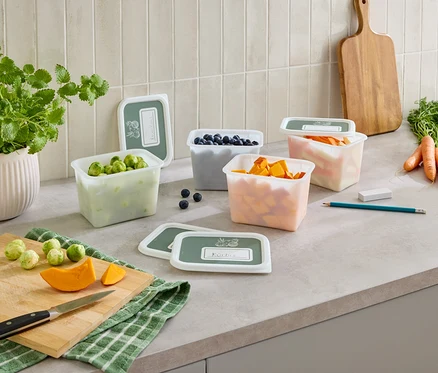 Sur un comptoir de cuisine, plusieurs récipients transparents avec couvercles, remplis de choux de Bruxelles, de myrtilles, de courge et de carottes.