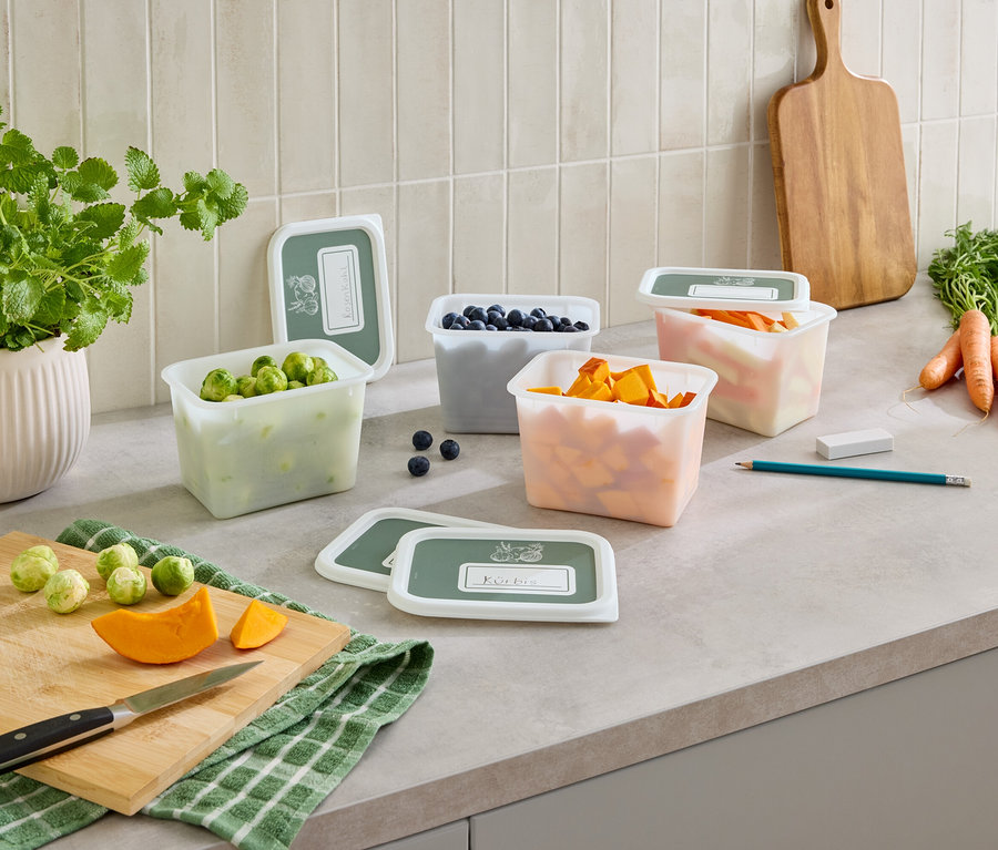 Sur un comptoir de cuisine, plusieurs récipients transparents avec couvercles, remplis de choux de Bruxelles, de myrtilles, de courge et de carottes.