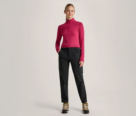 Une femme aux cheveux blonds attachés en queue de cheval se tient debout, les mains dans les poches. Elle porte un haut rose à manches longues avec un col zippé, un pantalon noir et des chaussures de randonnée marron.