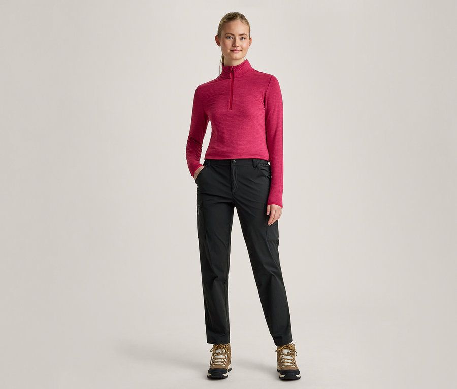 Une femme aux cheveux blonds attachés en queue de cheval se tient debout, les mains dans les poches. Elle porte un haut rose à manches longues avec un col zippé, un pantalon noir et des chaussures de randonnée marron.