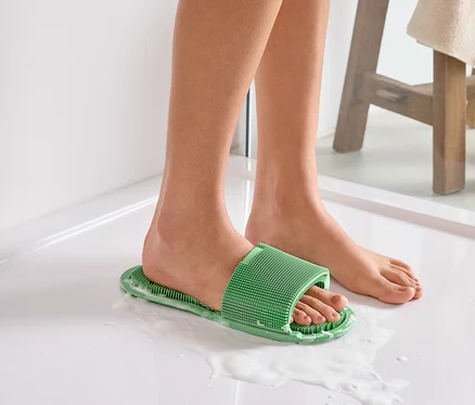 Gros plan des pieds, dont un portant une sandale de bain verte à picots, sur un sol de douche blanc savonneux.