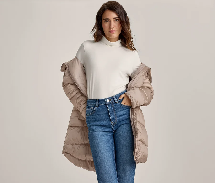 Femme aux cheveux foncés portant un col roulé clair, un jean bleu et une doudoune beige drapée sur ses épaules, une main dans sa poche, regardant l'appareil photo.