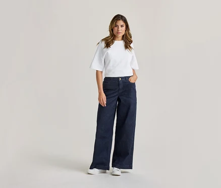 Femme debout, vêtue d'un t-shirt blanc à manches courtes, d'un jean large bleu foncé et de baskets blanches. Sa main droite est dans sa poche.