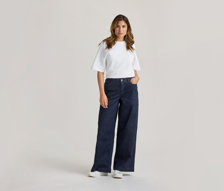 Femme debout, vêtue d'un t-shirt blanc à manches courtes, d'un jean large bleu foncé et de baskets blanches. Sa main droite est dans sa poche.