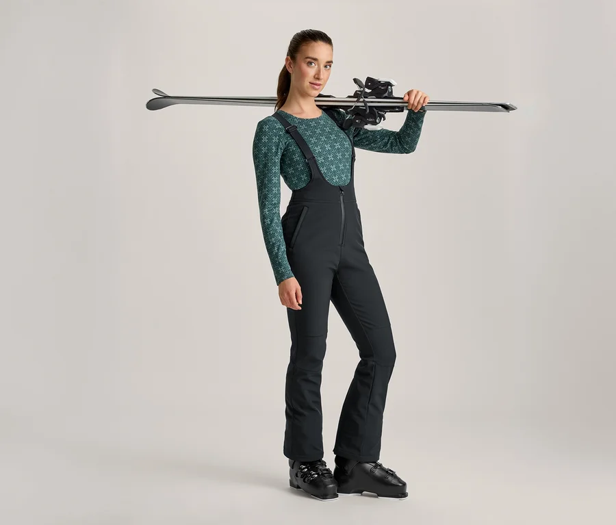 Femme en haut à manches longues vert à motifs, pantalon de ski noir et chaussures de ski, portant des skis sur son épaule.