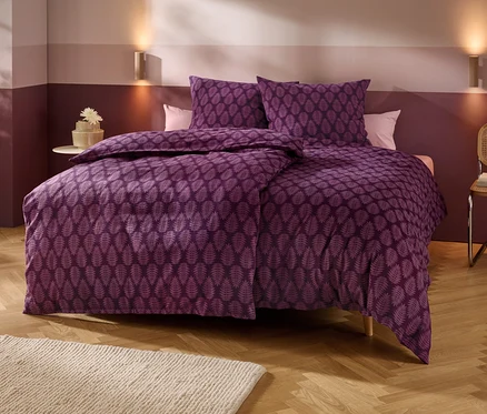 Un lit avec une parure de lit violette à motif de feuilles se trouve dans une chambre aux murs bicolores, au parquet en bois et à un tapis clair. Deux appliques murales éclairent la zone au-dessus du lit. À gauche, une petite table d'appoint avec une fleur, à droite, une chaise avec des livres.