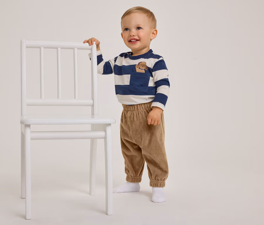 Un tout-petit souriant, vêtu d'un t-shirt rayé bleu et blanc et d'un pantalon en velours côtelé marron, se tient à côté d'une chaise blanche en la tenant.