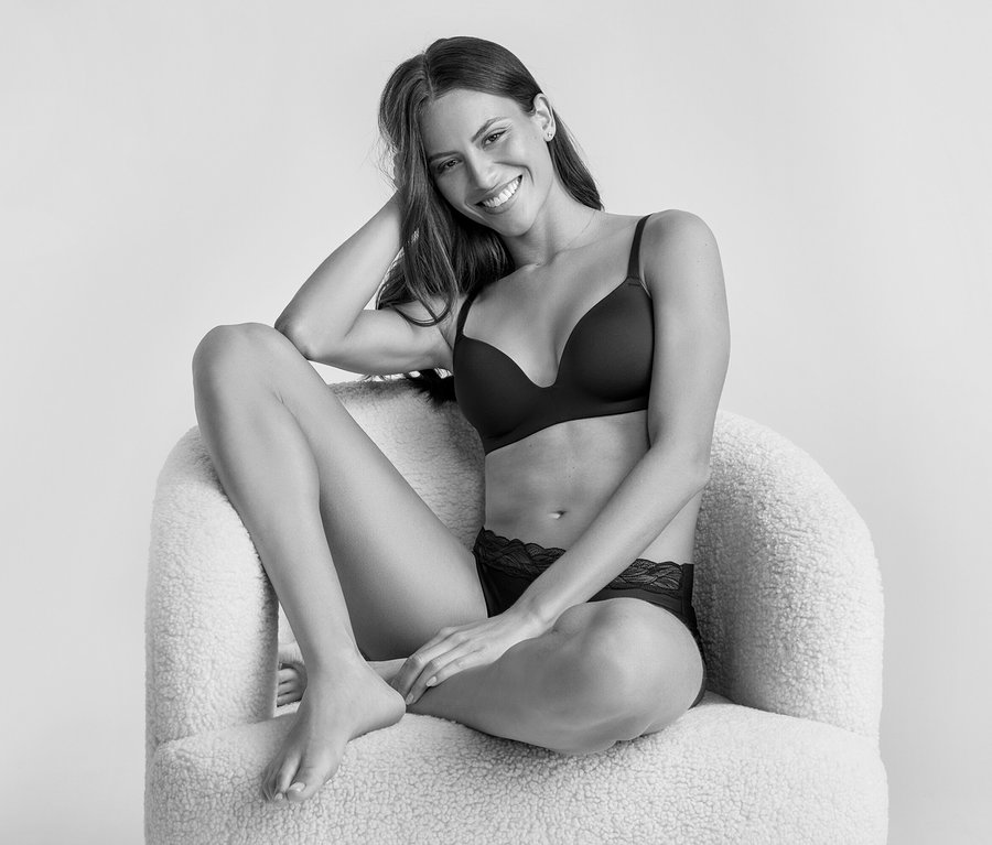 Une femme souriante aux longs cheveux foncés est assise sur un fauteuil moelleux, vêtue d'un soutien-gorge à bonnets souples noir et d'une culotte en dentelle noire. Elle regarde l'appareil photo, un bras posé sur sa tête.