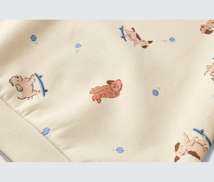 Gros plan d'un tissu beige clair avec un motif de chiens de dessin animé et de balles bleues. Certains chiens sont sur des skateboards, d'autres sont allongés. Un poignet côtelé est visible en bas à gauche.