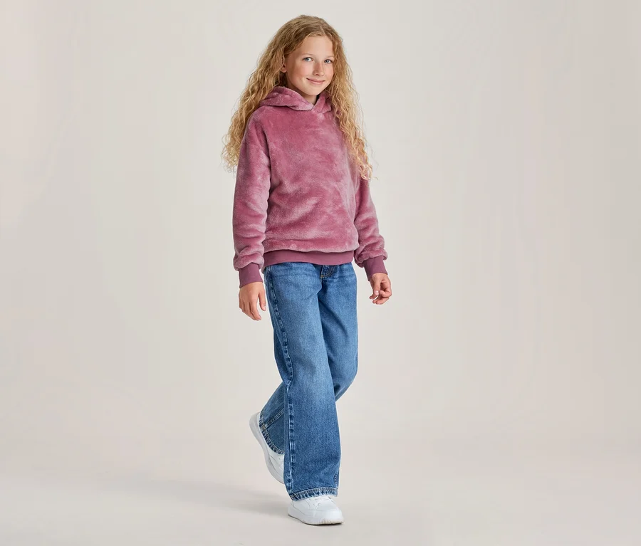 Une jeune fille aux longs cheveux blonds bouclés sourit en marchant, portant un sweat à capuche rose duveteux, un jean bleu et des baskets blanches.