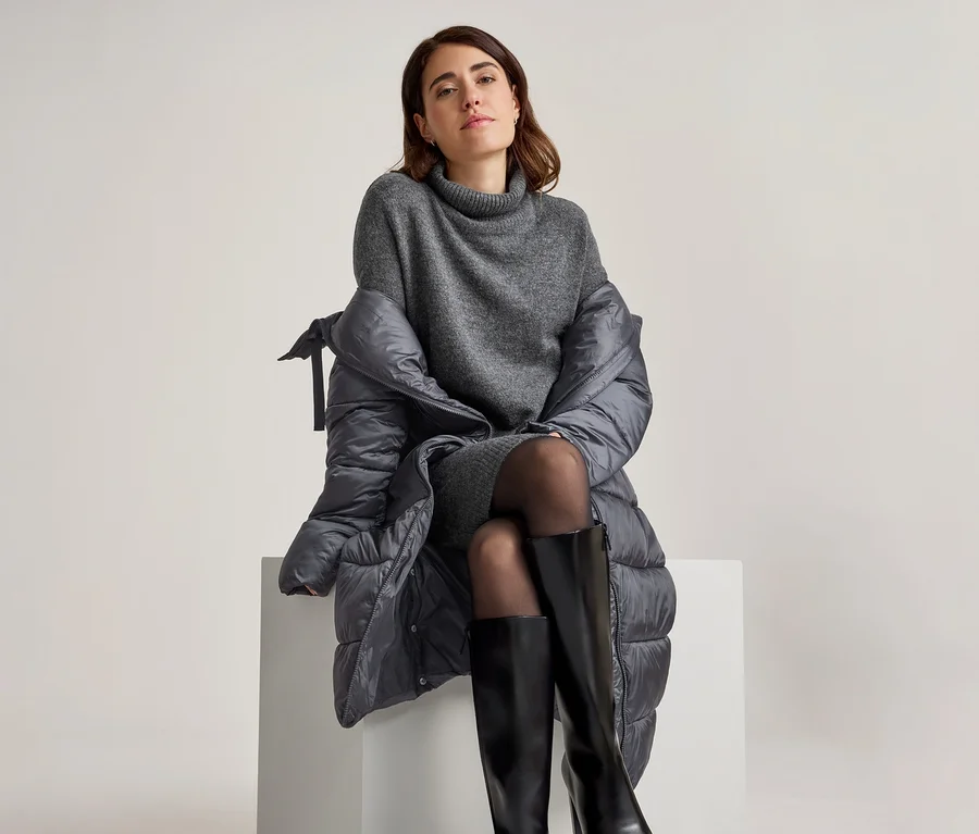 Femme assise sur un bloc blanc, portant une robe pull grise à col roulé, des collants noirs et des bottes hautes noires, avec un manteau matelassé gris foncé drapé sur ses épaules.