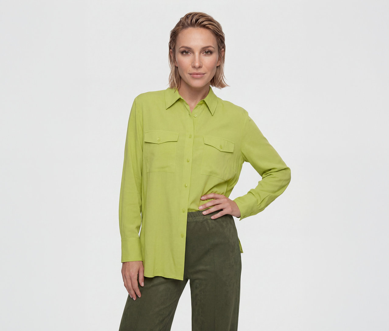 Femme aux cheveux courts portant une chemise vert citron à manches longues avec deux poches poitrine et un pantalon vert foncé, posant une main sur la hanche.