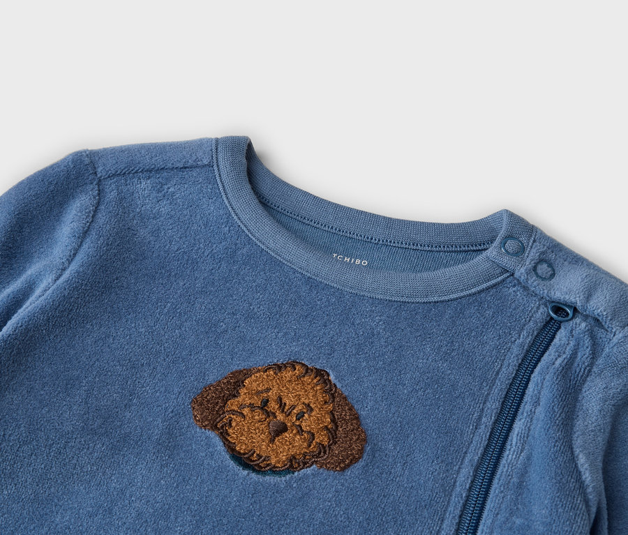 Grenouillère bébé bleue en tissu doux, avec un visage de chien brodé marron, des boutons-pression à l'épaule gauche et une fermeture éclair bleue diagonale.