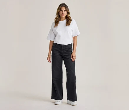Femme debout, portant un t-shirt blanc à manches courtes, un jean large gris foncé et des baskets blanches.
