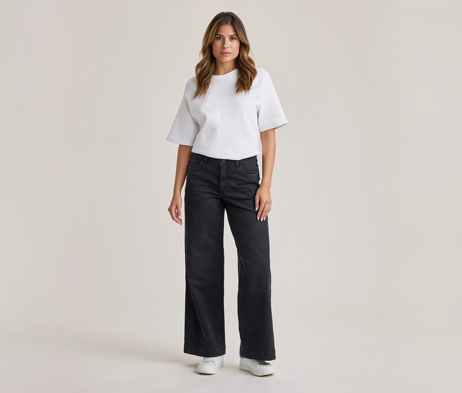 Femme debout, portant un t-shirt blanc à manches courtes, un jean large gris foncé et des baskets blanches.