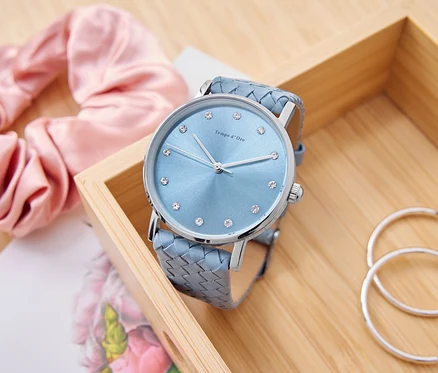 Une montre bleu clair avec des strass est posée dans une boîte en bois, à côté d'un chouchou rose, d'un bloc-notes et de créoles argentées.