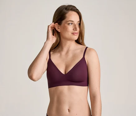 Une femme aux longs cheveux bruns et aux taches de rousseur porte un soutien-gorge bordeaux et touche son oreille.