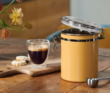 Café dans un verre, biscuits sur une planche en bois et un récipient à café jaune ouvert avec des grains, une cuillère doseuse, sur une table en bois avec des fleurs.