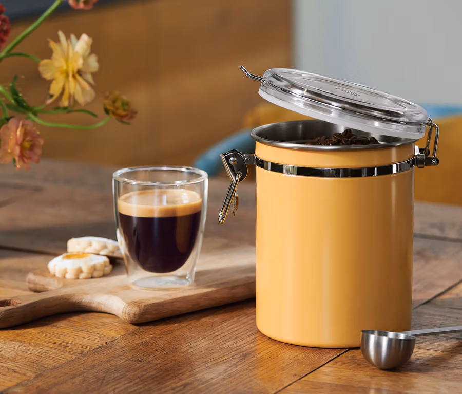 Café dans un verre, biscuits sur une planche en bois et un récipient à café jaune ouvert avec des grains, une cuillère doseuse, sur une table en bois avec des fleurs.