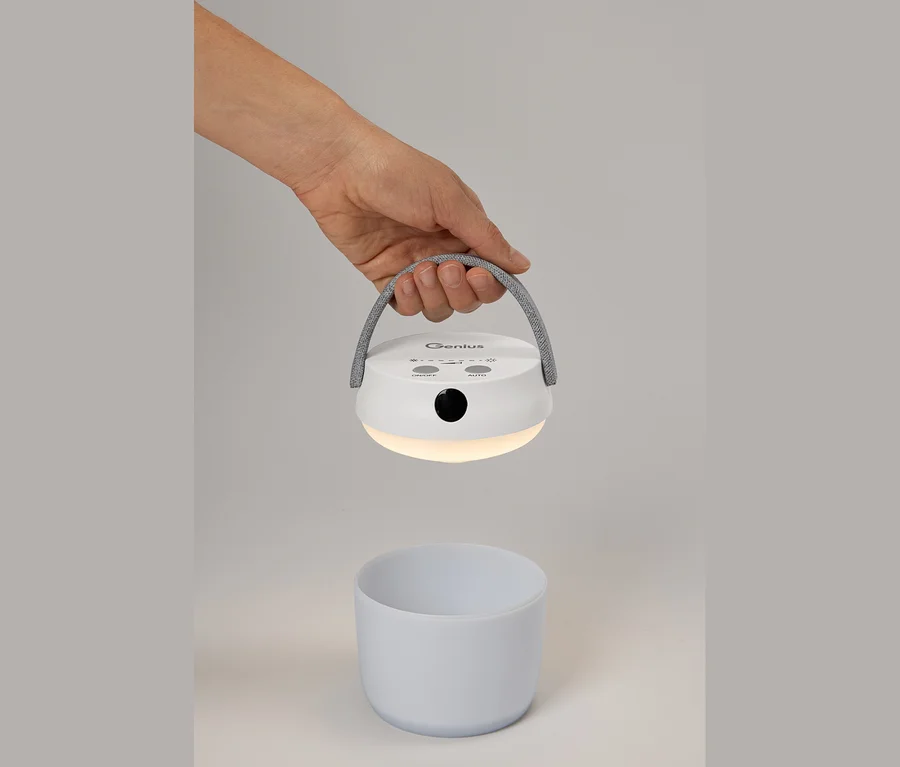 Une main tient une lampe LED multifonction blanche et allumée avec détecteur de mouvement et poignée en tissu gris, au-dessus d'une tasse blanche.
